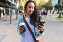 Asian woman using smartphone and holding takeaway coffee in the street. independent young woman out and about in the city. — Stock Photo