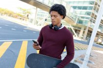 Африканський американський чоловік у місті користується смартфоном і тримає скейтборд. Цифровий кочівник у русі, десь у місті.. — стокове фото