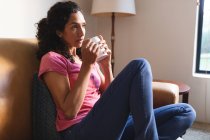 Mixed race woman sitting on sofa and drinking coffee. domestic lifestyle and spending quality time at home. — Stock Photo