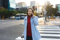 Retrato de mujer asiática usando smartphone y cruzando calle con maleta. mujer joven independiente fuera y alrededor de la ciudad. - foto de stock
