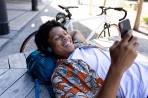 Hombre afroamericano en la ciudad mintiendo y usando un teléfono inteligente. nómada digital sobre la marcha, fuera y alrededor de la ciudad. - foto de stock