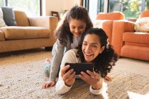 Raza mixta madre e hija acostadas en la alfombra, usando un teléfono inteligente. estilo de vida doméstico y pasar tiempo de calidad en casa. - foto de stock