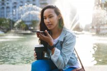 Mujer asiática hablando en smartphone y sosteniendo café para llevar en el soleado parque. mujer joven independiente fuera y alrededor de la ciudad. - foto de stock