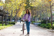 Retrato de mujer asiática sonriente rodando bicicleta y mirando a la cámara en el soleado parque. mujer joven independiente fuera y alrededor de la ciudad. - foto de stock