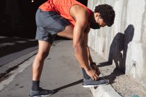 Fit african american man exercising in city wearing face mask, tying shoes. fitness and active urban outdoor lifestyle during coronavirus covid 19 pandemic. — Stock Photo