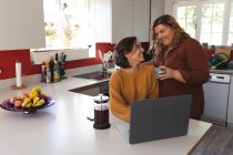 Lesbisches Paar lächelt und benutzt Laptop in der Küche. häuslicher Lebensstil, Freizeit zu Hause verbringen. — Stockfoto