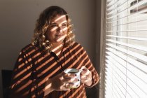 Mulher branca sorrindo e bebendo café ao lado da janela. estilo de vida doméstico, passar o tempo livre em casa. — Fotografia de Stock