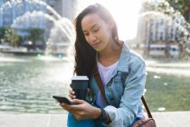 Donna asiatica utilizzando smartphone e tenendo il caffè da asporto nel parco soleggiato. indipendente giovane donna in giro per la città. — Foto stock