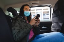 Asiatin mit Gesichtsmaske sitzt im Taxi und benutzt Smartphone. Unabhängige junge Frau während der Coronavirus-Pandemie 19 in der Stadt unterwegs. — Stockfoto