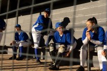 Diverse Baseballspielerinnen sitzen auf Bänken in der Sonne und unterhalten sich und warten darauf, zu spielen. Baseballmannschaft, Sporttraining, Zusammenhalt und Engagement. — Stockfoto