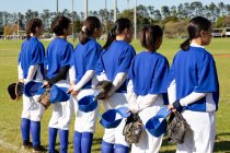 Разнообразная группа женщин-бейсболистов, стоящих на поле с руками за спиной перед игрой. женская бейсбольная команда, спортивные тренировки, сплоченность и приверженность. — стоковое фото