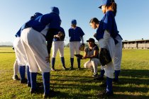 Разнообразная группа женщин-бейсболистов, стоящих в обнимку вокруг тренера на поле. женская бейсбольная команда, спортивная подготовка и тактика игры. — стоковое фото