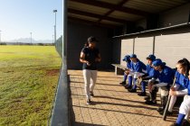 Grupo diverso de jogadoras de beisebol sentadas no banco, ouvindo treinadoras antes do jogo. time de beisebol feminino, treinamento esportivo e táticas de jogo. — Fotografia de Stock