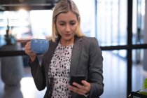 Mulher de negócios caucasiana usando smartphone e bebendo caneca de café. trabalhando em negócios em um escritório moderno. — Fotografia de Stock