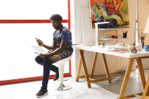 Pintor afroamericano en el trabajo usando un smartphone en un estudio de arte. creación e inspiración en un estudio de pintura de artistas. - foto de stock
