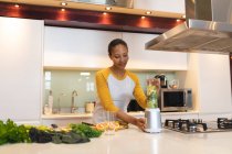Souriante femme métissée dans la cuisine préparant boisson santé. mode de vie domestique, profiter du temps libre à la maison. — Photo de stock