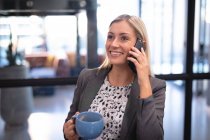 Mujer de negocios caucásica usando teléfono inteligente y beber taza de café. trabajar en un negocio en una oficina moderna. - foto de stock