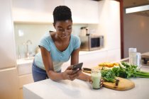 Lächelnde Afroamerikanerin in der Küche, die Gesundheitsgetränk trinkt und Smartphone benutzt. häuslicher Lebensstil, Freizeit zu Hause genießen. — Stockfoto