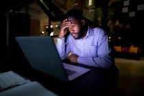 Hombre de negocios afroamericano trabajando de noche, sentado en el escritorio y usando un portátil. trabajar hasta tarde en los negocios en una oficina moderna. - foto de stock