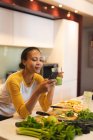 Mujer sonriente de raza mixta en la cocina bebiendo bebidas saludables y usando un teléfono inteligente. estilo de vida doméstico, disfrutando del tiempo libre en casa. - foto de stock
