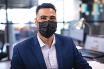 Porträt eines Geschäftsmannes mit gemischter Rasse, der Gesichtsmaske trägt und in die Kamera blickt. Arbeit in einem modernen Büro während der Coronavirus-Pandemie 19. — Stockfoto