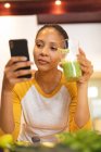 Femme souriante métissée dans la cuisine boire une boisson santé et en utilisant un smartphone. mode de vie domestique, profiter du temps libre à la maison. — Photo de stock