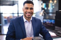 Портрет усміхненого змішаного гоночного бізнесмена, який п'є каву і дивиться на камеру. робота в бізнесі в сучасному офісі . — стокове фото