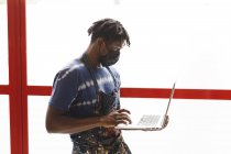 Pintor masculino afroamericano usando mascarilla facial usando laptop en estudio de arte. creación e inspiración en un estudio de pintura de artistas durante coronavirus covid 19 pandemia. - foto de stock