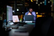 Un uomo d'affari afroamericano che lavora di notte, seduto alla scrivania e usa un portatile. lavorare fino a tardi negli affari in un ufficio moderno. — Foto stock
