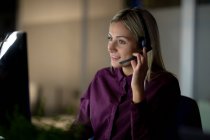 Mujer de negocios caucásica que trabaja de noche con auriculares. trabajar hasta tarde en los negocios en una oficina moderna. - foto de stock