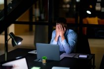 Empresario de carrera mixta trabajando de noche, sentado en el escritorio y usando un portátil. trabajar hasta tarde en los negocios en una oficina moderna. - foto de stock