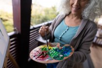 Mulher caucasiana sênior feliz em pé na varanda ensolarada e pintura. estilo de vida aposentadoria, passar o tempo sozinho em casa. — Fotografia de Stock