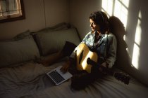 Gemischte Rassenfrau spielt Gitarre und benutzt Laptop im sonnigen Schlafzimmer. Gesunder Lebensstil, Freizeit zu Hause genießen. — Stockfoto