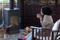 Fröhliche Mischlingsfrau im Wohnzimmer am Kamin sitzend, Tasse haltend und Kaffee trinkend. Auszeit zu Hause. . — Stockfoto