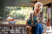 Relajante mujer mayor caucásica en la cocina con teléfono inteligente y beber café. estilo de vida de jubilación, pasar tiempo solo en casa. - foto de stock