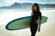 Портрет змішаної раси жінки, яка тримає дошку для серфінгу в сонячний день на пляжі. здоровий спосіб життя, насолоджуючись дозвіллям на відкритому повітрі . — стокове фото