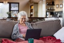 Donna caucasica anziana seduta sul divano e che utilizza il computer portatile nel moderno soggiorno. stile di vita di pensione, trascorrere del tempo da solo a casa. — Foto stock