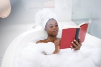Happy african american woman in bathroom relaxing in bath reading book. domestic lifestyle, enjoying self care leisure time at home. — Stock Photo