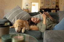 Усміхнена біла жінка у вітальні, лежить на дивані зі своїм домашнім собакою, використовуючи смартфон. домашній спосіб життя, насолоджуючись дозвіллям вдома . — стокове фото