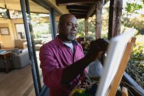 Homem americano africano sênior na varanda ensolarada pintando um quadro. estilo de vida aposentadoria, passar o tempo sozinho em casa. — Fotografia de Stock