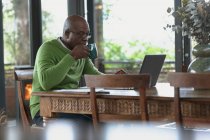 Uomo anziano afroamericano che si trova e lavora sul computer portatile nel moderno soggiorno. stile di vita di pensione, trascorrere del tempo da solo a casa. — Foto stock