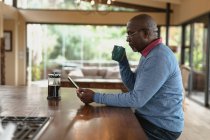 Hombre afroamericano mayor bebiendo café y usando tableta en la cocina moderna. estilo de vida de jubilación, pasar tiempo solo en casa. - foto de stock