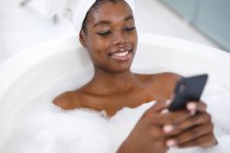 Усміхнена афро-американська жінка у ванній кімнаті, відпочиваючи у ванній, використовуючи смартфон. Домашній спосіб життя, дозвілля для себе вдома. — стокове фото
