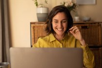 Donna caucasica sorridente in soggiorno, seduta al tavolo di lavoro, con il computer portatile che indossa cuffie. stile di vita domestico, lavoro a distanza da casa. — Foto stock