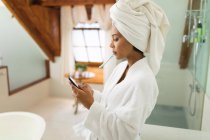 Змішана расова жінка у ванній кімнаті за допомогою смартфона та чищення зубів. домашній спосіб життя, насолоджуючись доглядом за дозвіллям вдома . — стокове фото