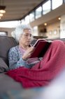 Mujer caucásica mayor sentada en el sofá y leyendo un libro en la sala de estar moderna. estilo de vida de jubilación, pasar tiempo solo en casa. - foto de stock