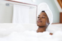 Sorrindo afro-americana em casa de banho relaxante em banho com os olhos fechados. estilo de vida doméstico, desfrutando de tempo de lazer auto-cuidado em casa. — Fotografia de Stock