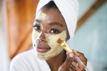 Ritratto di donna afroamericana sorridente in bagno applicando maschera viso di bellezza. stile di vita domestico, godendo di auto cura del tempo libero a casa. — Foto stock