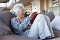 Feliz mujer caucásica mayor sentada en el sofá y leyendo el libro en la sala de estar moderna. estilo de vida de jubilación, pasar tiempo solo en casa. - foto de stock