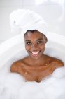 Ritratto di donna afroamericana sorridente in bagno, rilassante nella vasca da bagno, guardando la macchina fotografica. stile di vita domestico, godendo di auto cura del tempo libero a casa. — Foto stock